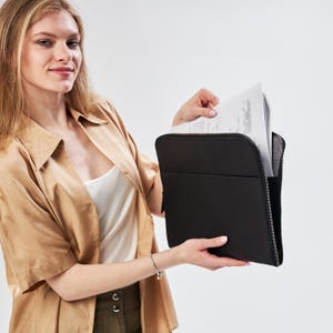 Könnte beinhalten: Eine Frau hält ein schwarzes Lederportfolio mit Reißverschluss. Das Portfolio ist geöffnet und enthält weiße Papiere.