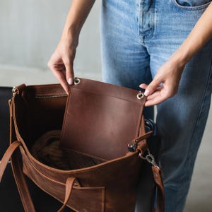 Puede incluir: Un bolso tote de cuero marr&oacute;n con cierre de cremallera y un bolsillo interior grande. El bolso est&aacute; abierto y el interior es visible, mostrando un forro de tela marr&oacute;n.