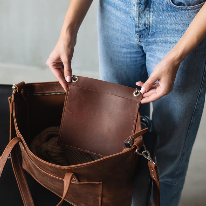 Puede incluir: Un bolso tote de cuero marr&oacute;n con cierre de cremallera y un bolsillo interior grande. El bolso est&aacute; abierto y el interior es visible, mostrando un forro de tela marr&oacute;n.
