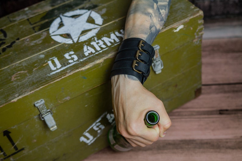 May include: Black leather wrist cuff with three buckles. The cuff is worn on a wrist with a tattoo. The background includes a green wooden box with the text "US ARMY" and a white star.