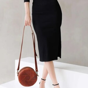 May include: A brown leather round crossbody bag with a geometric design. The bag is worn by a person wearing a black dress and black heels.