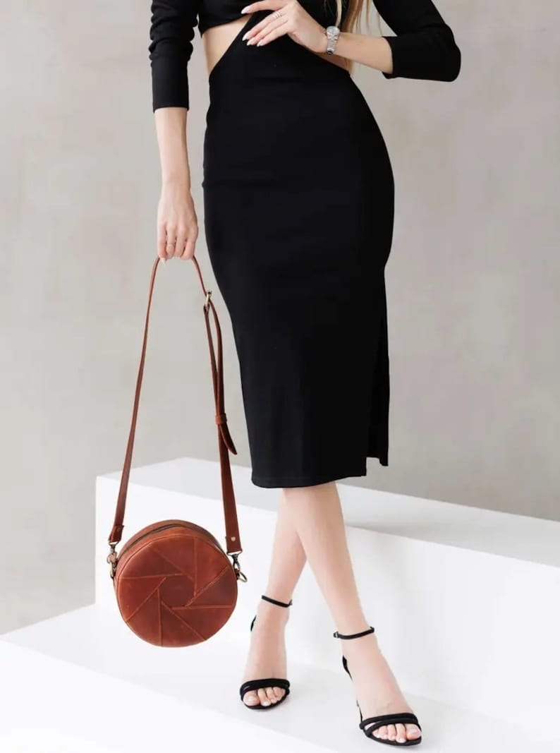 May include: A brown leather round crossbody bag with a geometric design. The bag is worn by a person wearing a black dress and black heels.