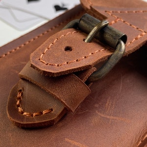 May include: Close-up of a brown leather card holder with a brass buckle. The buckle is secured with a stitched leather strap.