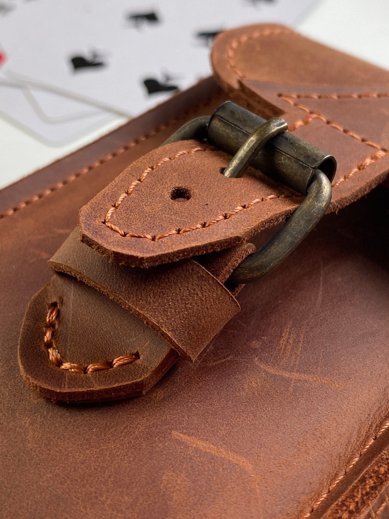 May include: Close-up of a brown leather card holder with a brass buckle. The buckle is secured with a stitched leather strap.