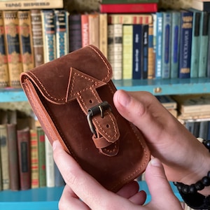 May include: A brown leather phone case with a buckle closure. The case has a stitched arrow design on the front.