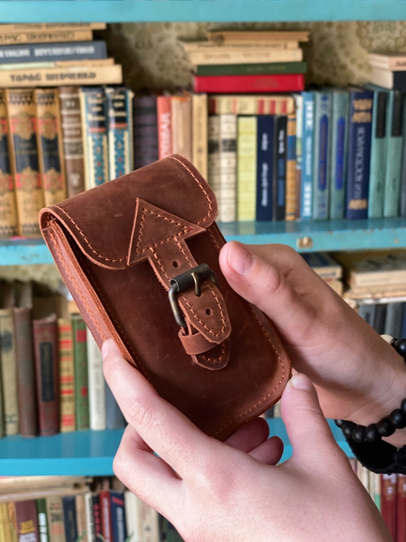 May include: A brown leather phone case with a buckle closure. The case has a stitched arrow design on the front.