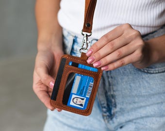May include: A brown leather ID holder with a clear window showing a blue ID card. The holder has a matching brown lanyard and a silver clasp. The holder also has a slot for cards. The ID holder is held by a person.