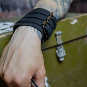 May include: Black leather wrist cuff with multiple horizontal straps and a brass-coloured buckle closure. The cuff is worn on a wrist with visible tattoos. The background includes a green box and a brick wall.