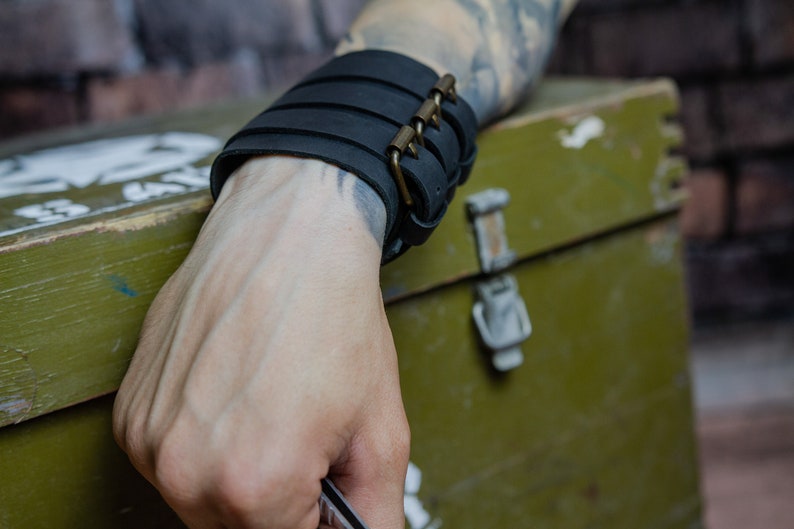 May include: Black leather wrist cuff with multiple horizontal straps and a brass-coloured buckle closure. The cuff is worn on a wrist with visible tattoos. The background includes a green box and a brick wall.