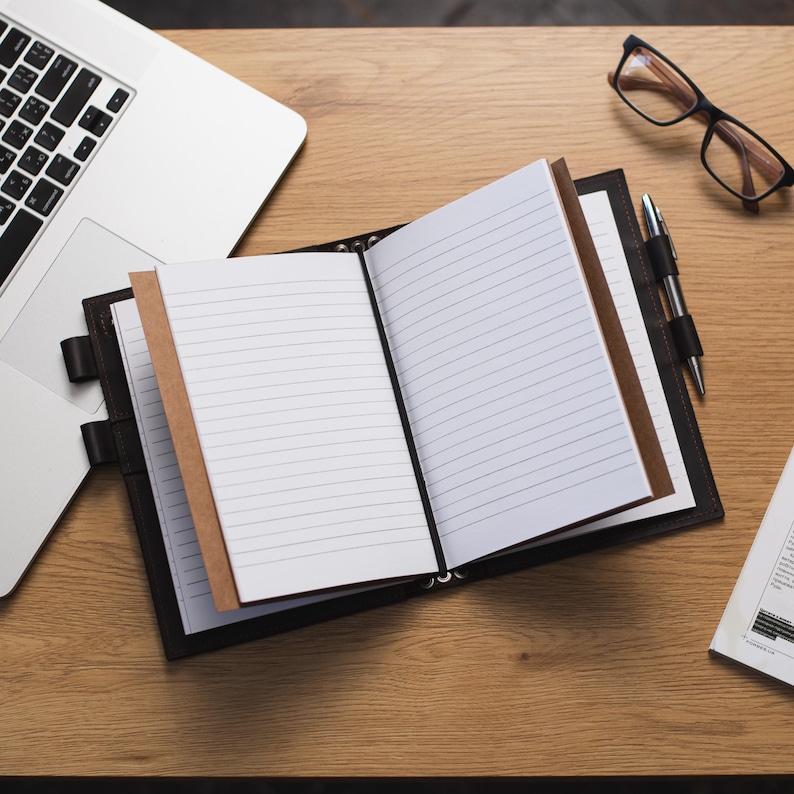 May include: A black leather bound notebook with lined pages is open on a wooden desk. A laptop computer, a pen, and eyeglasses are also on the desk.