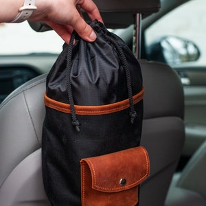 Op de afbeelding: Een zwarte canvas tas met een bruine leren rand en een klein bruin leren zakje. De tas hangt aan de hoofdsteun van een auto.