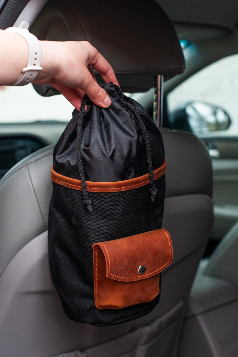 Op de afbeelding: Een zwarte canvas tas met een bruine leren rand en een klein bruin leren zakje. De tas hangt aan de hoofdsteun van een auto.