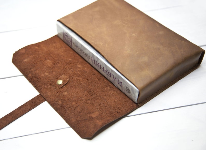 May include: A brown leather book cover with a strap and a snap closure. The cover is open to reveal a book titled "НИНДЖУ И ПРОКЛЯТИЯ".