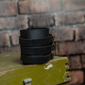 May include: A stack of black leather wrist cuffs with brass-coloured buckles. The cuffs are stacked on top of each other, set against a brick wall and a green wooden box.