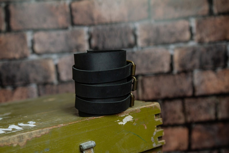 May include: A stack of black leather wrist cuffs with brass-coloured buckles. The cuffs are stacked on top of each other, set against a brick wall and a green wooden box.