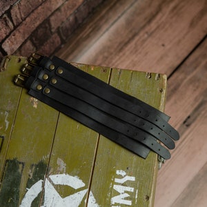 May include: Several black leather wrist cuffs with brass buckles and rivets are arranged on a distressed green wooden box. The cuffs are stacked, highlighting their design. The background includes a brick wall and wooden planks.