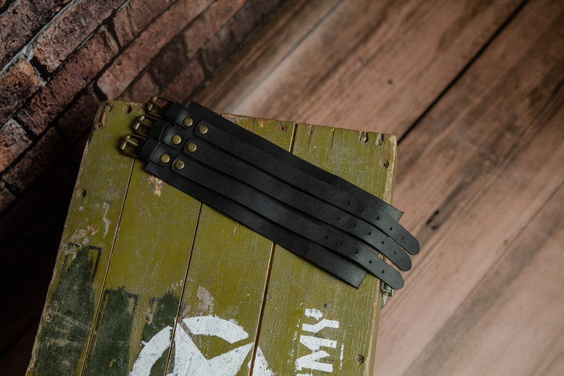 May include: Several black leather wrist cuffs with brass buckles and rivets are arranged on a distressed green wooden box. The cuffs are stacked, highlighting their design. The background includes a brick wall and wooden planks.