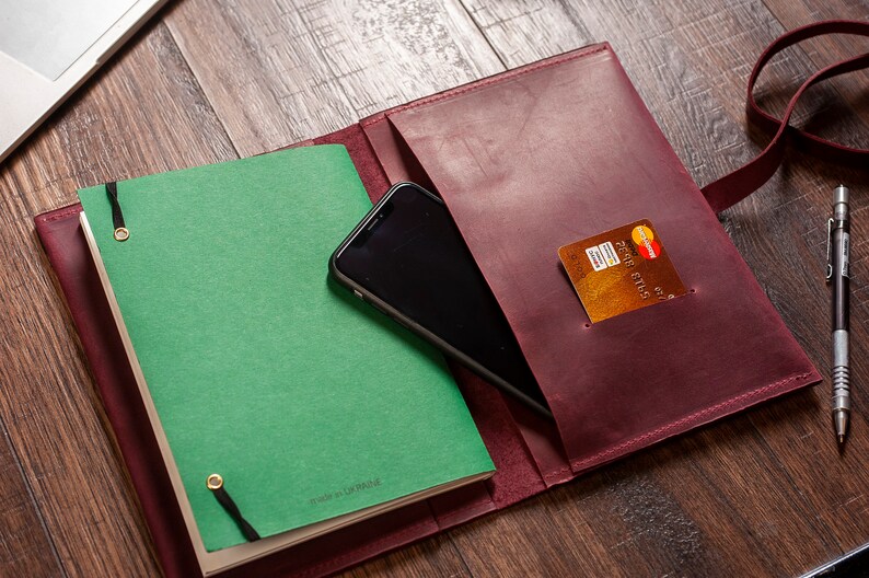 May include: A burgundy leather journal with a green notebook inside. The journal has a pocket on the outside that holds a credit card and a smartphone. The journal also has a strap and a pen.