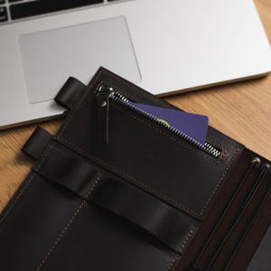 May include: A brown leather notebook with a zippered pocket and a card inside. The notebook has multiple slots for pens or other items.