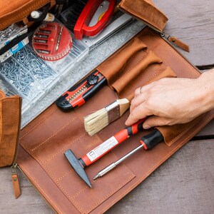 Pu&ograve; includere: Un rotolo portautensili in pelle marrone con vari strumenti, tra cui un martello, un cacciavite, un pennello e un coltellino multiuso. Il rotolo &egrave; aperto e disposto piatto su una superficie di legno.