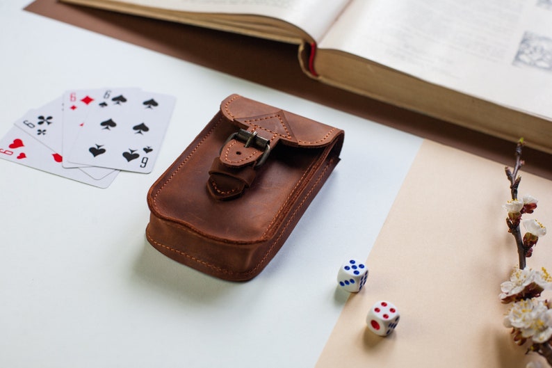 May include: A brown leather pouch with a buckle closure, playing cards, and two dice on a white and beige surface.