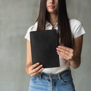 May include: A woman holds a brown leather notebook with a pen sticking out of the top. The notebook is closed and has a simple design.