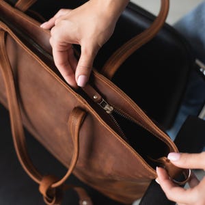 Puede incluir: Un bolso tote de cuero marr&oacute;n con cierre de cremallera. El bolso tiene un asa superior y una correa larga para el hombro.