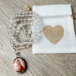 May include: A clear quartz crystal mala necklace with a silver pendant featuring a colorful image of a deity. The necklace is displayed on a white fabric pouch with a burlap heart.