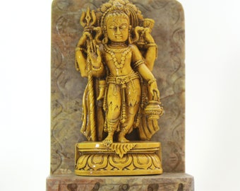 May include: A golden statue of a Hindu deity standing on a stone base. The deity is holding a trident and a pot in their hands. The statue is carved in detail and has a serene expression.