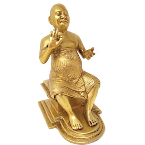 May include: A gold-colored metal statue of a seated figure with a rounded face and a patterned garment. The figure is sitting on a decorative base with intricate details.