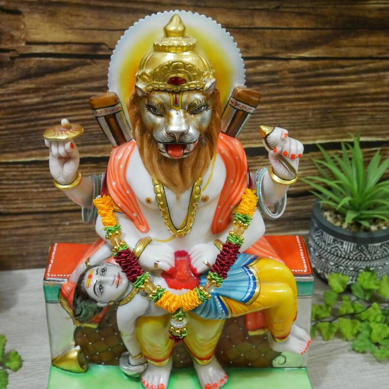 May include: A white marble statue of a Hindu deity with a lion head, sitting on a throne with a smaller figure lying at their feet. The deity is adorned with gold and red accents, and the smaller figure is wearing a blue and yellow garment.
