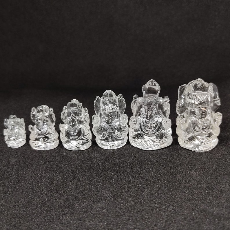 May include: A set of seven clear crystal Ganesha statues of varying sizes. The statues are all in the same pose, with the god Ganesha sitting with his trunk raised and his hands in a mudra. The statues are all made of clear crystal and have a smooth, polished finish.