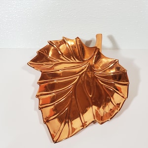 May include: A copper-colored leaf-shaped decorative dish. The dish has detailed veining and a stem. The metallic finish reflects light, creating highlights and shadows. The leaf dish is a decorative accent piece.