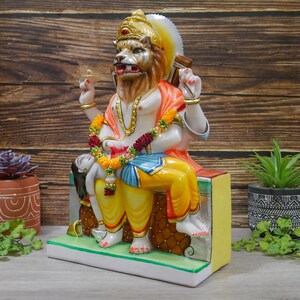 May include: A colorful statue of a deity with a lion head, sitting on a throne with a reclining figure. The statue is made of marble and is painted in vibrant colors, including red, yellow, blue, and green.