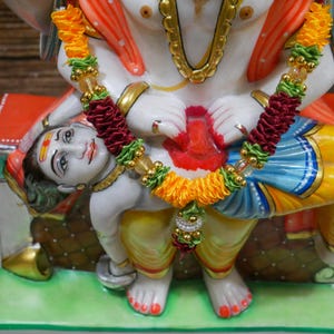 May include: A white marble statue of a deity with a red, yellow, and blue robe, holding a smaller white marble statue of a deity with a yellow, red, and blue robe. Both statues are adorned with a garland of yellow, green, and red fabric.