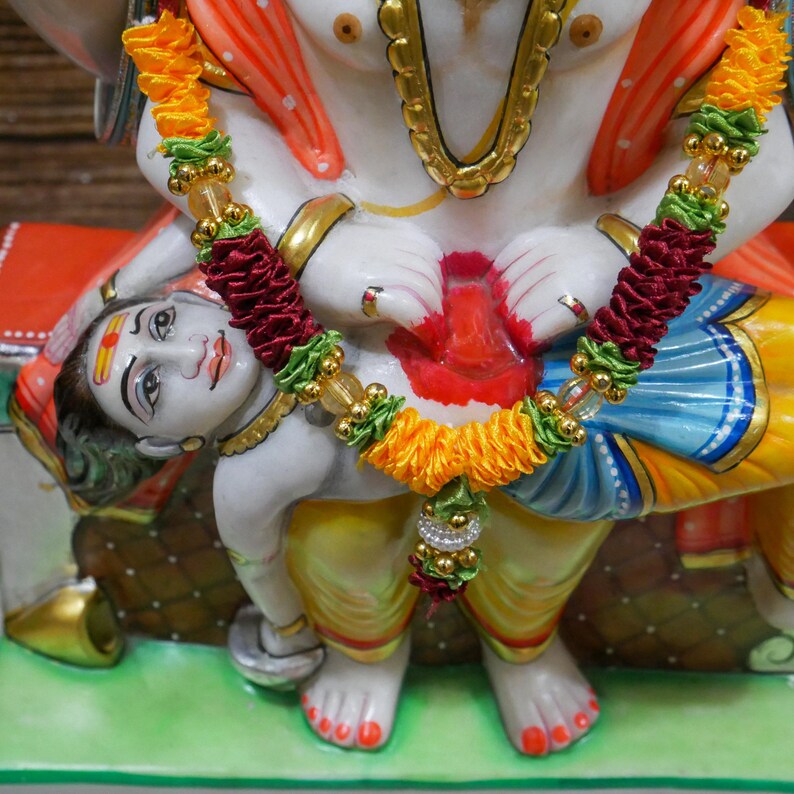 May include: A white marble statue of a deity with a red, yellow, and blue robe, holding a smaller white marble statue of a deity with a yellow, red, and blue robe. Both statues are adorned with a garland of yellow, green, and red fabric.