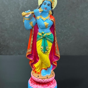 May include: A vibrant statue of a blue-skinned deity playing a flute. The figure wears a yellow dhoti, green sash, and red cape. The statue is adorned with gold jewelry and stands on a lotus flower base, approximately 6 inches tall.