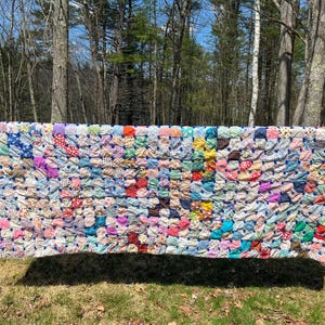 May include: A colorful patchwork quilt, displayed outdoors. The quilt features a variety of fabric patterns and colors, including blue, red, yellow, and purple. The quilt is rectangular and appears to be handmade, with a textured surface.