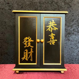 May include: Black cabinet with gold trim and two doors. Gold Chinese characters are on the doors. The cabinet has two gold knobs and sits on four small gold feet. The cabinet is on a red surface.