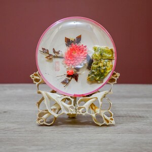 May include: A decorative, round, clear glass piece with a pink rim, featuring a floral design with a red flower and green accents. The glass piece is displayed on a carved, off-white stand, showcasing an artistic display.