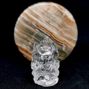 May include: A clear crystal Ganesha statue, a Hindu deity, sitting in a meditative pose with its trunk curled up. The statue is carved with intricate details and is set against a background of a round, multi-colored stone.