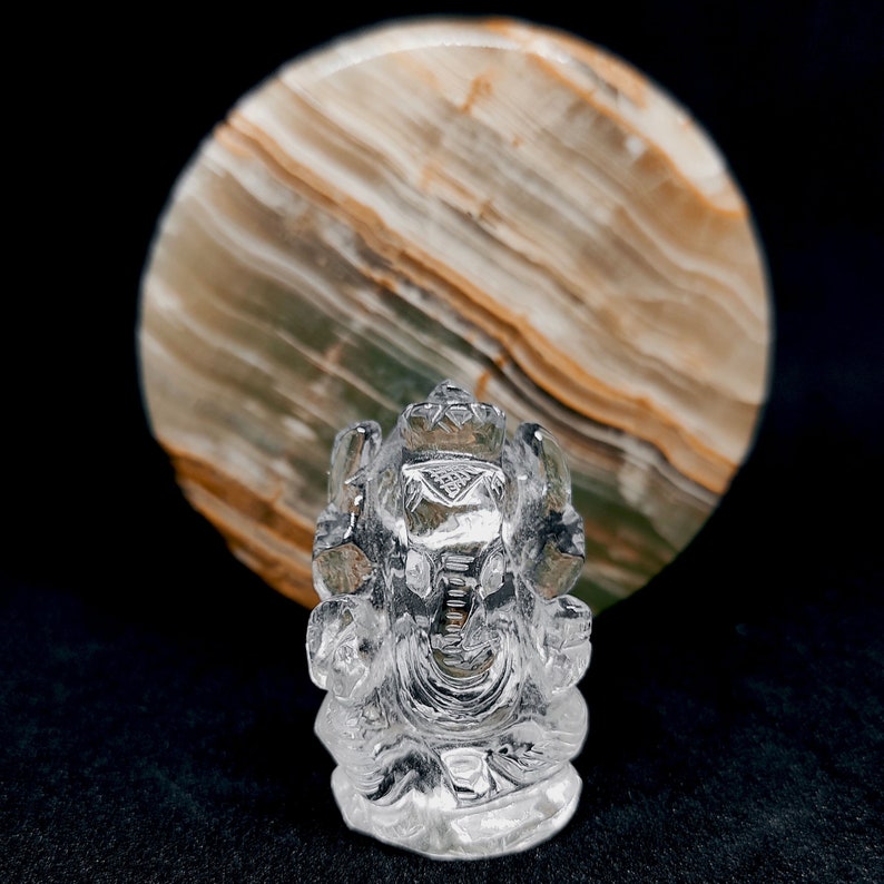 May include: A clear crystal Ganesha statue, a Hindu deity, sitting in a meditative pose with its trunk curled up. The statue is carved with intricate details and is set against a background of a round, multi-colored stone.