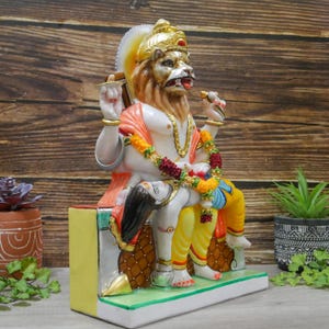May include: A white marble statue of a Hindu deity with a lion head, sitting on a throne with a female figure at his feet. The statue is decorated with colorful paint and intricate details.