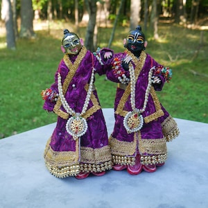 May include: Two standing deity figurines dressed in matching purple robes with gold trim and intricate embroidery. Each figure wears a long pearl necklace with a central pendant, beaded wrist cuffs, and has a distinct facial complexion, one gold and one black.