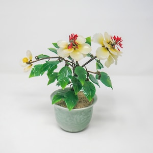 May include: A decorative potted plant with white and yellow flowers with red accents, green leaves, and a light green pot. The plant is a detailed, artistic representation of a flowering plant.