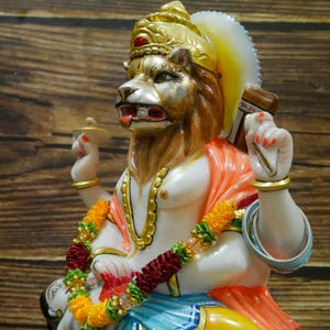 May include: A white marble statue of a deity with a lion head, wearing a gold crown and orange and white clothing. The statue is decorated with gold paint and has a garland of yellow and red flowers around its neck.