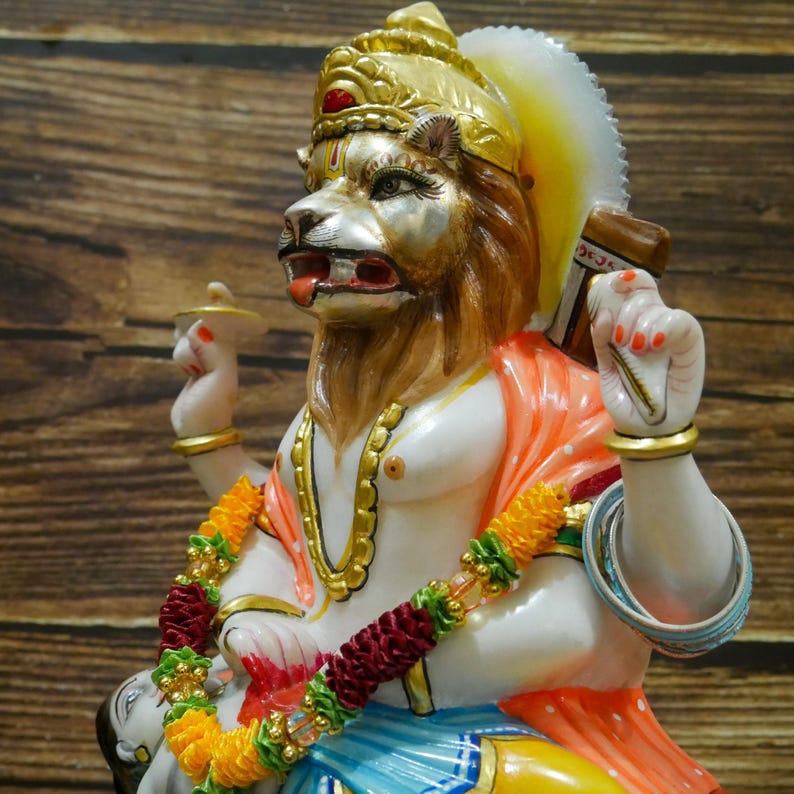 May include: A white marble statue of a deity with a lion head, wearing a gold crown and orange and white clothing. The statue is decorated with gold paint and has a garland of yellow and red flowers around its neck.