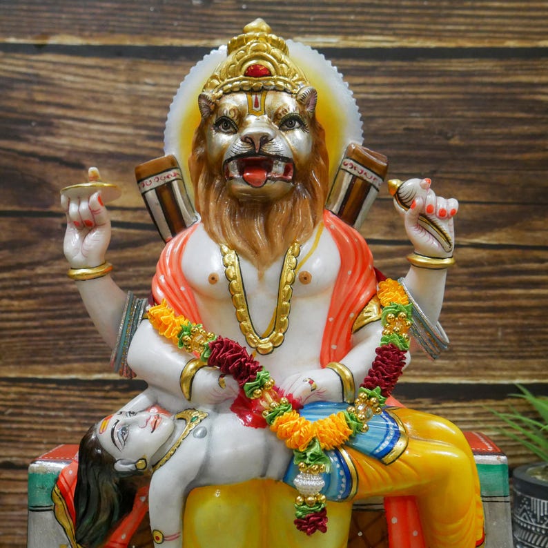 May include: A white marble statue of a Hindu deity with a lion head, sitting on a throne with a figure lying at their feet. The deity is adorned with gold jewelry and a red and orange sash. The figure at their feet is wearing a blue and yellow sash.