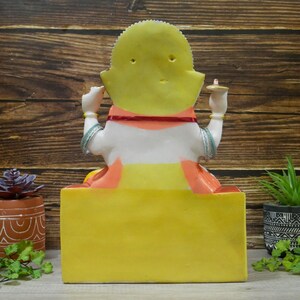 May include: A white and yellow statue of a seated goddess with a red and orange sash. The statue is on a yellow base and has a small, yellow halo behind the head.