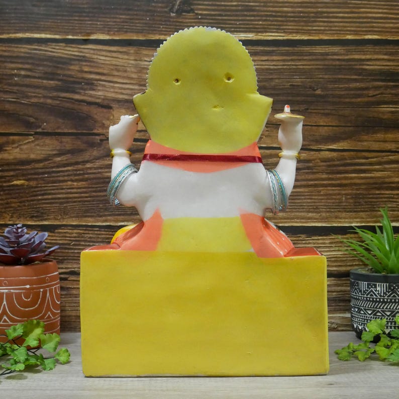 May include: A white and yellow statue of a seated goddess with a red and orange sash. The statue is on a yellow base and has a small, yellow halo behind the head.